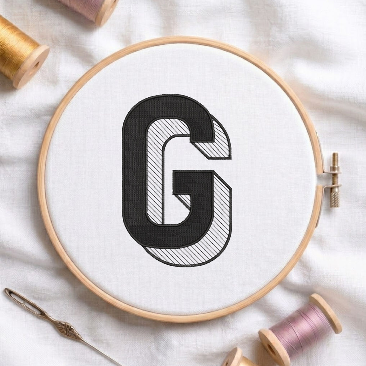 A wooden embroidery hoop holding white fabric with a large black embroidered letter G, surrounded by spools of thread and a needle on a white fabric background.