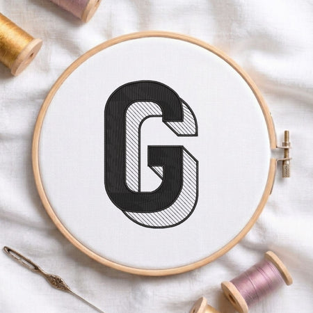 A wooden embroidery hoop holding white fabric with a large black embroidered letter G, surrounded by spools of thread and a needle on a white fabric background.