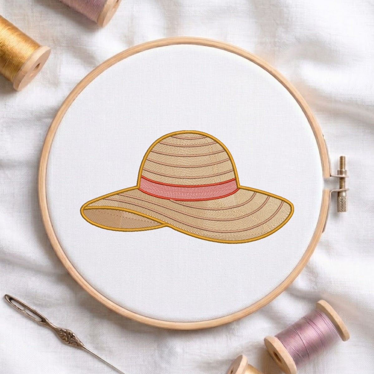 A circular embroidery hoop holds white fabric with a stitched sun hat design in beige, pink, and gold thread. A needle and spools of thread are arranged around it on a white fabric background.