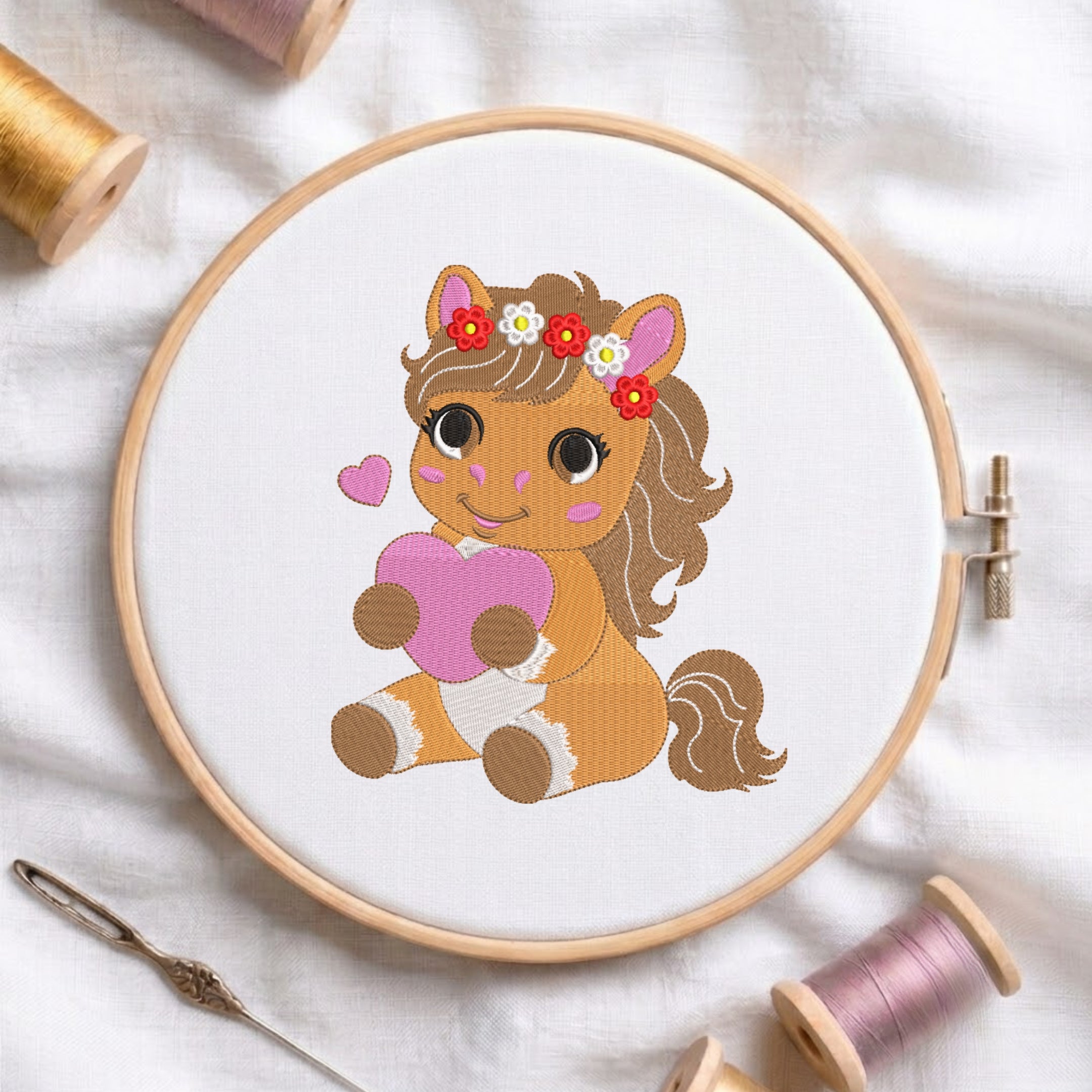 A circular embroidery hoop displays a cartoon horse design with a pink heart and floral headband, surrounded by spools of thread and a needle on a white fabric background.