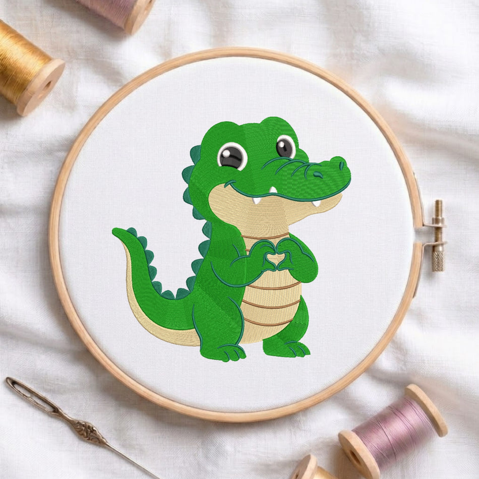 A white fabric stretched in a wooden embroidery hoop, featuring an embroidered bright green alligator with a tan belly, smiling eyes, and its hands forming a small heart shape.