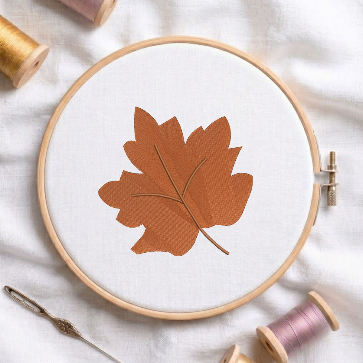 A circular embroidery hoop holding white fabric with a stitched brown leaf design, surrounded by spools of thread and a needle on a white fabric background.