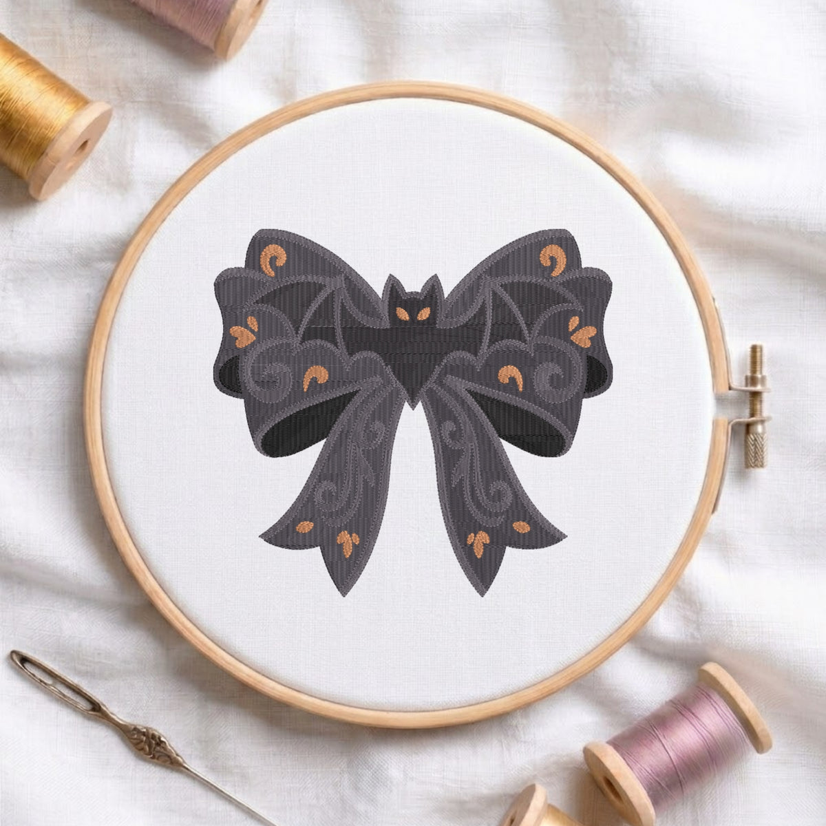 A white fabric stretched in a wooden embroidery hoop, featuring an embroidered design of a dark patterned bow adorned with wing details and centered with a vampire bat.