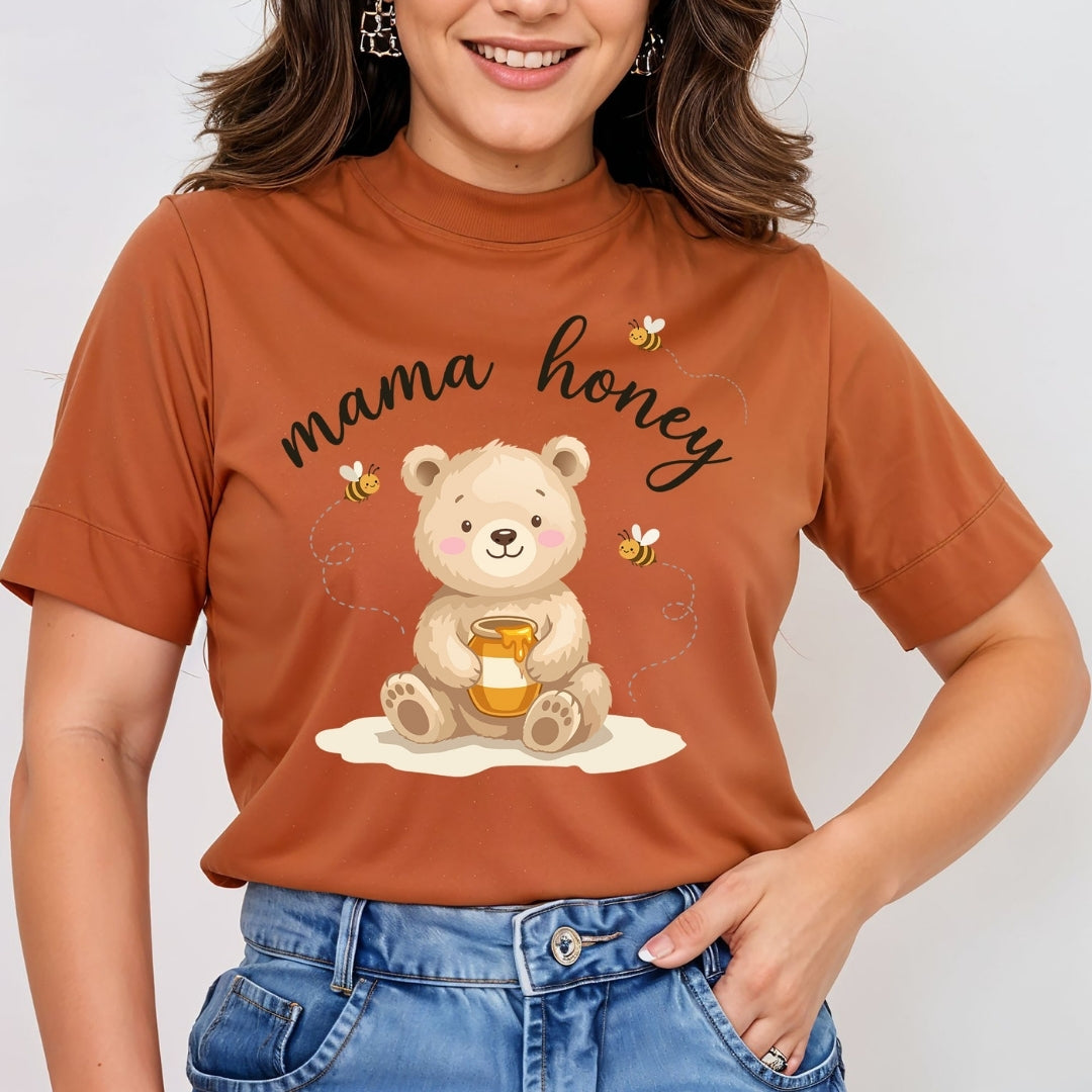 A woman wearing a rust-orange short-sleeve t-shirt with a cartoon graphic of a teddy bear holding a honey pot and the words 'mama honey' in black script, paired with blue jeans.