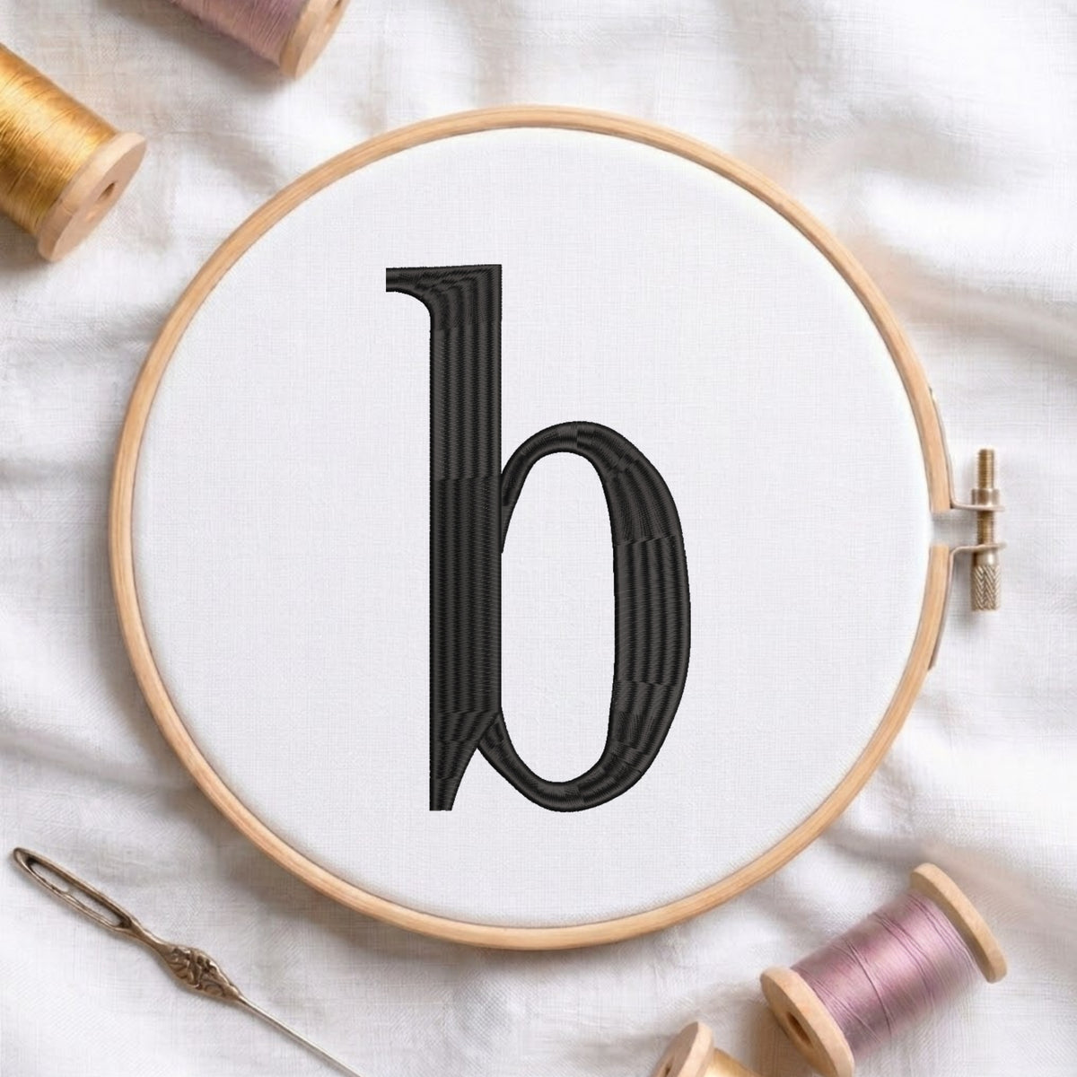 A wooden embroidery hoop holding white fabric with a large black letter 'b' stitched in the center, styled on a white fabric background with spools of thread and a needle.