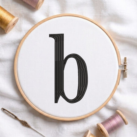 A wooden embroidery hoop holding white fabric with a large black letter 'b' stitched in the center, styled on a white fabric background with spools of thread and a needle.