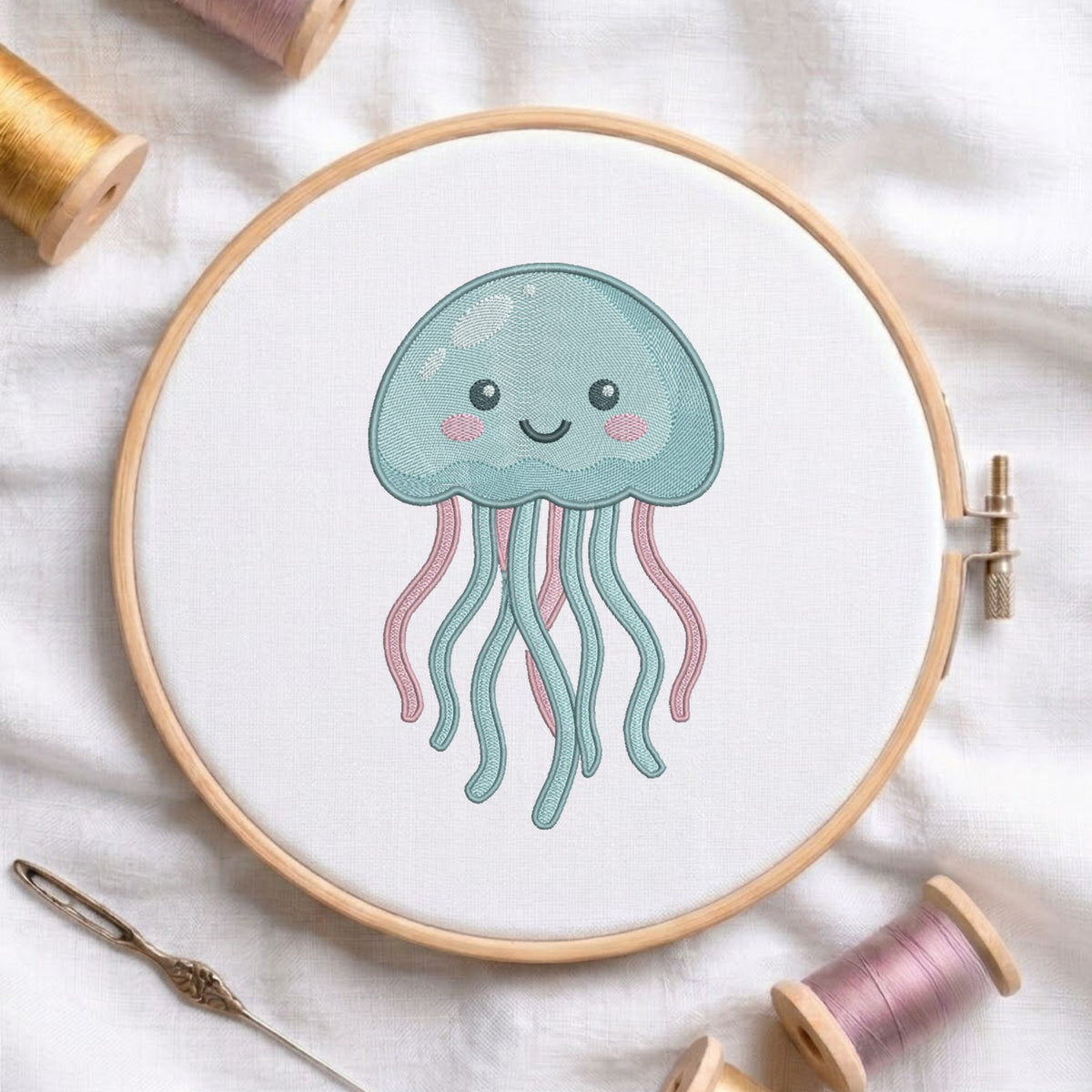 A wooden embroidery hoop holds white fabric with a cute embroidered jellyfish design in light blue and pink, surrounded by spools of thread and a needle on a white fabric background.