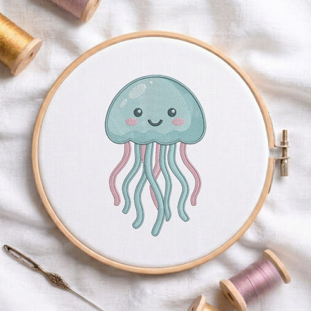 A wooden embroidery hoop holds white fabric with a cute embroidered jellyfish design in light blue and pink, surrounded by spools of thread and a needle on a white fabric background.