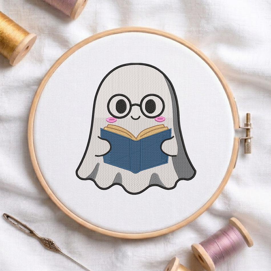 A circular embroidery hoop holding a white fabric with a cute ghost character wearing glasses and reading a blue book. The hoop is styled on a white fabric background with spools of thread and a needle.