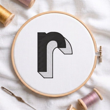 A wooden embroidery hoop holding white fabric with a black-and-white letter 'r' design, surrounded by spools of thread and a needle on a white fabric background.