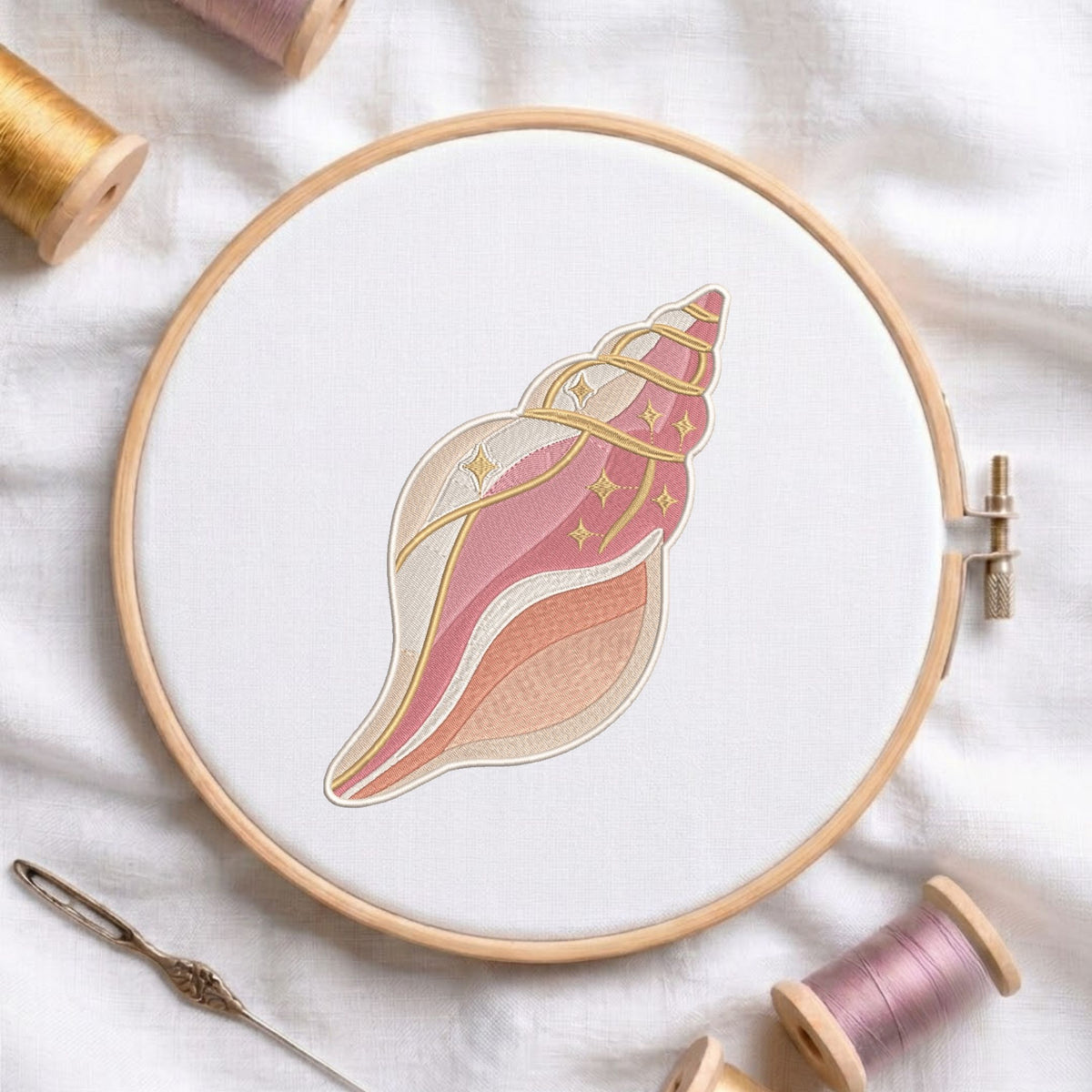 Pink and beige conch seashell machine embroidery design displayed inside a round wooden hoop on white fabric alongside thread spools and a needle.