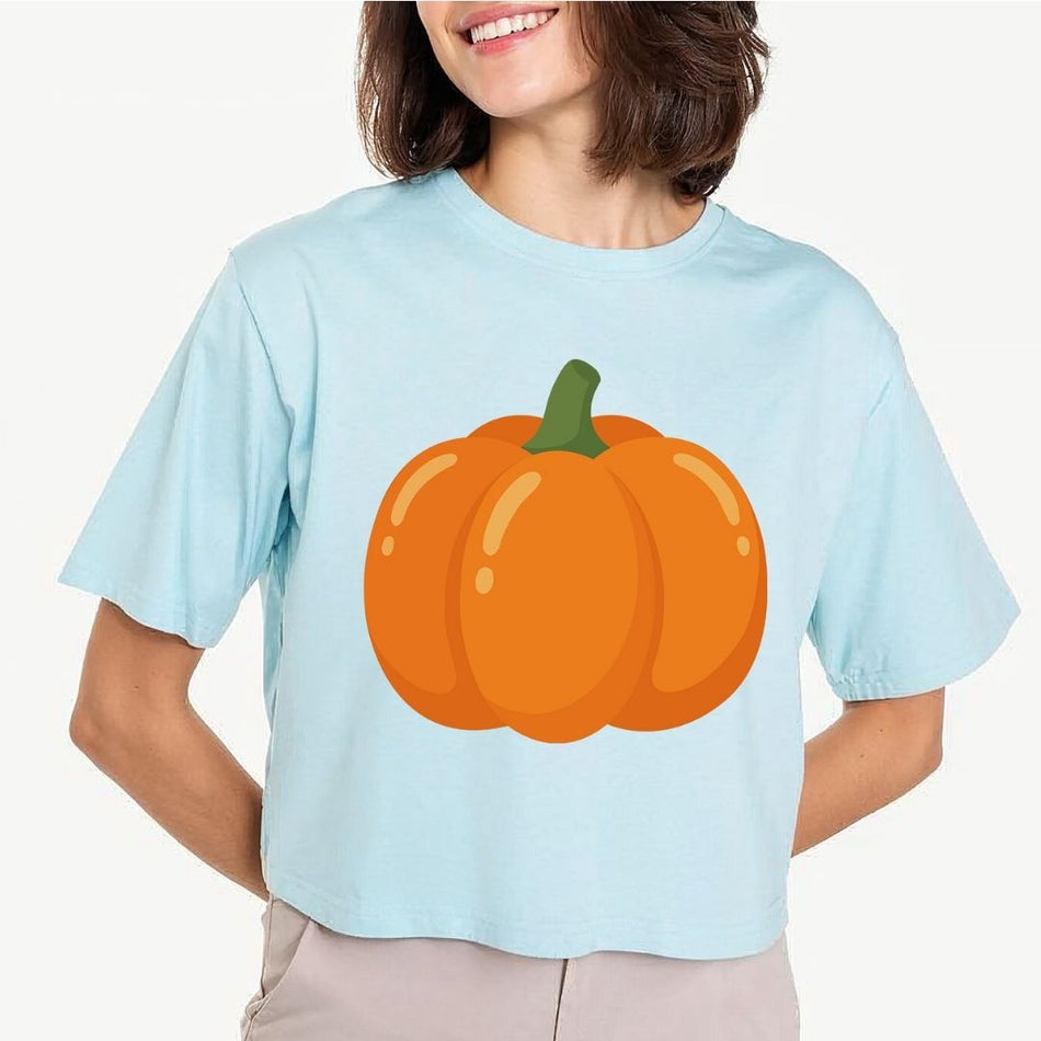 A woman wearing a light blue cropped t-shirt with a large orange pumpkin graphic on the front, paired with beige pants, against a plain light gray background.