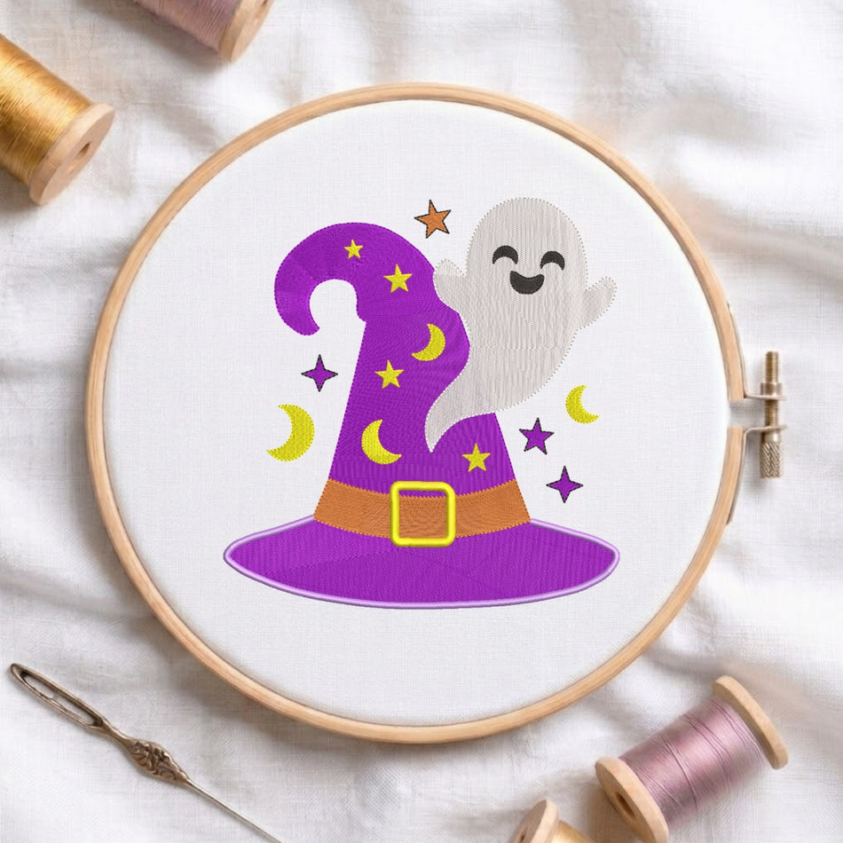 A circular embroidery hoop holding white fabric with a colorful witch hat and ghost design. The design includes a purple hat with yellow crescent moons and stars, a white smiling ghost, and a brown belt with a yellow buckle.