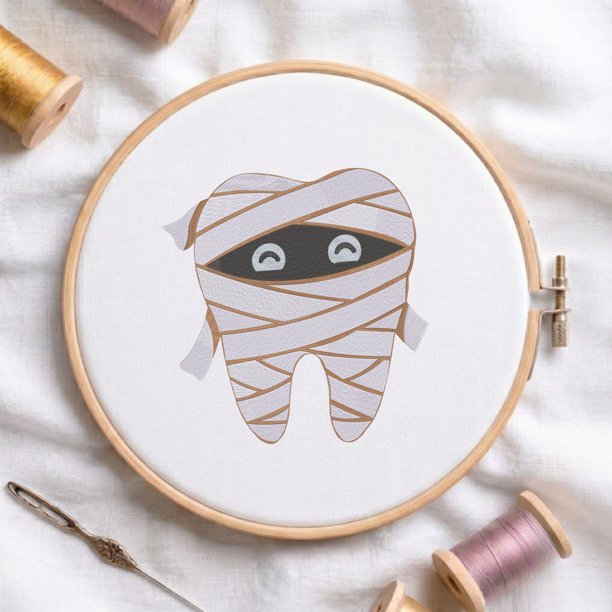 Cute mummy tooth Halloween dental machine embroidery design shown in a wooden embroidery hoop on white fabric with thread spools and a needle   