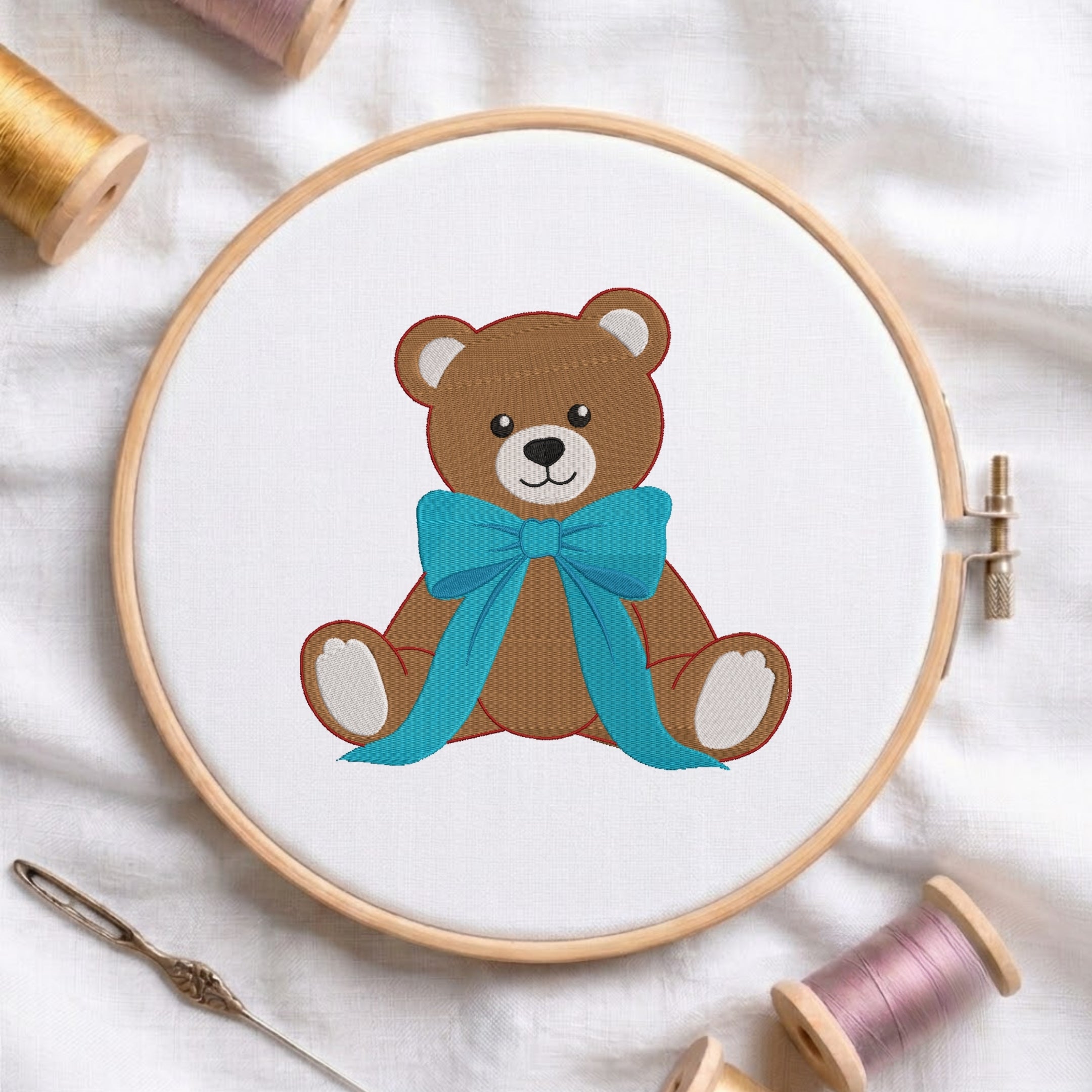 A wooden embroidery hoop holds white fabric with a stitched brown teddy bear wearing a bright turquoise bow. The hoop is styled on a white fabric background with spools of thread, a needle, and a pin around it.