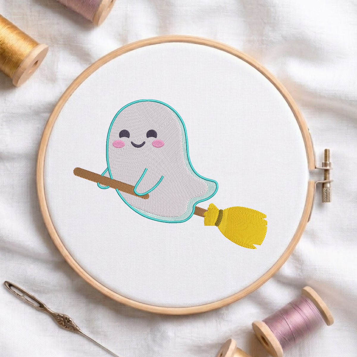 A circular embroidery hoop on white fabric holds a cute embroidered ghost holding a yellow broomstick. Spools of thread and a needle are arranged around it on a soft white fabric background.