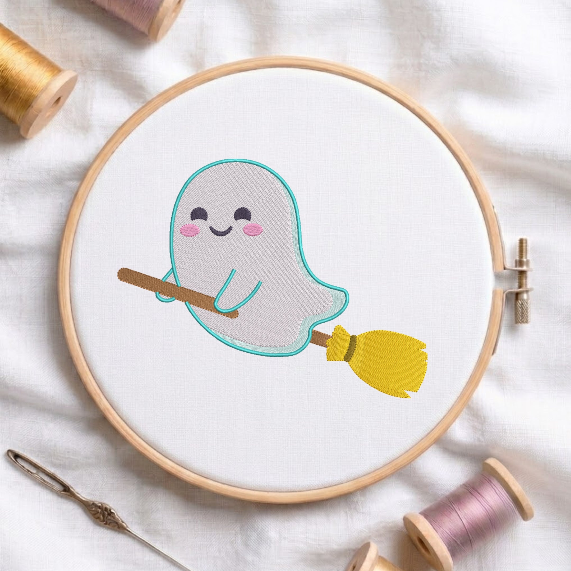 A circular embroidery hoop on white fabric holds a cute embroidered ghost holding a yellow broomstick. Spools of thread and a needle are arranged around it on a soft white fabric background.