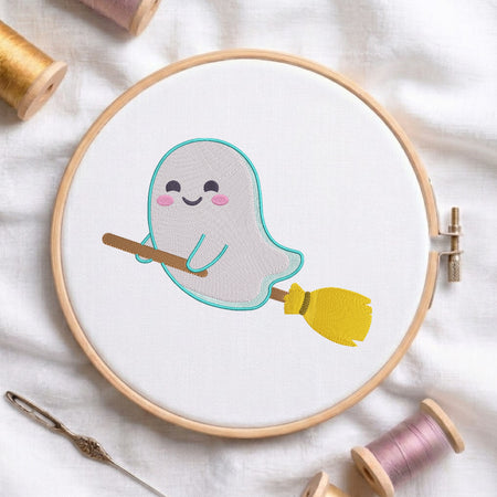 A circular embroidery hoop on white fabric holds a cute embroidered ghost holding a yellow broomstick. Spools of thread and a needle are arranged around it on a soft white fabric background.