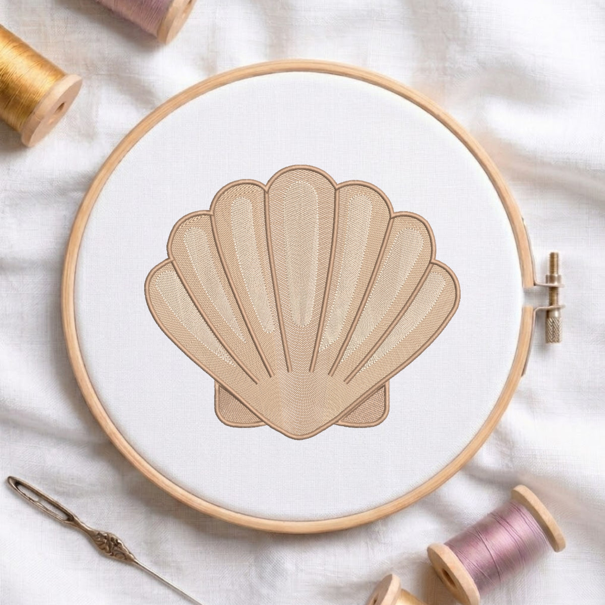 A shell-shaped embroidery design stitched in beige thread within a wooden embroidery hoop, surrounded by spools of thread and a needle on a white fabric background.