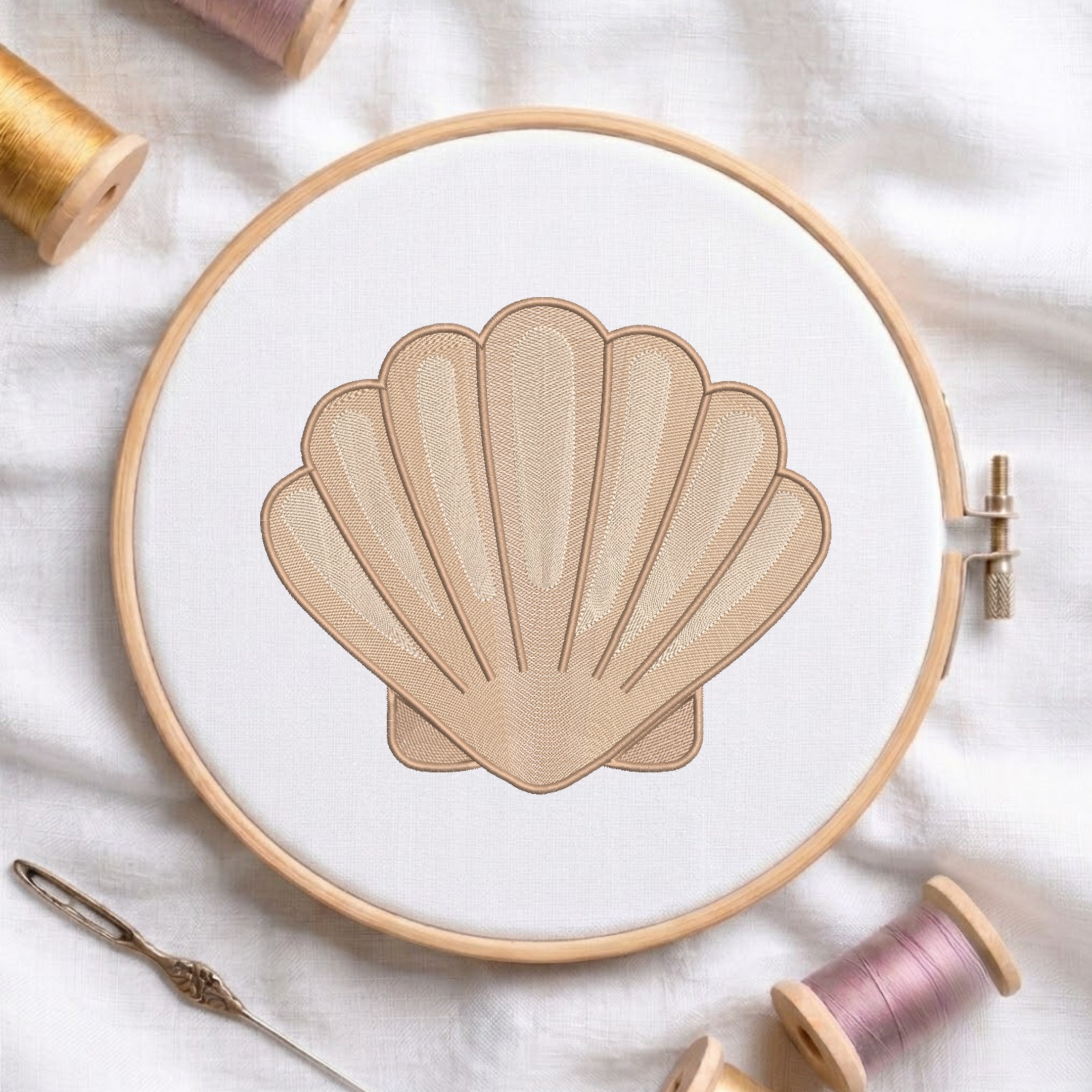A shell-shaped embroidery design stitched in beige thread within a wooden embroidery hoop, surrounded by spools of thread and a needle on a white fabric background.