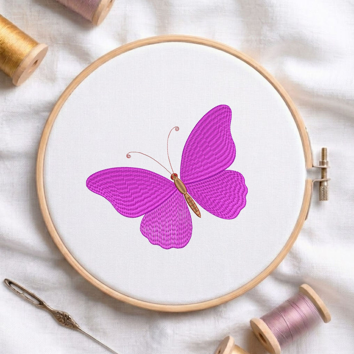 Vibrant purple butterfly machine embroidery design displayed inside a round wooden hoop on white fabric with thread spools and a needle.