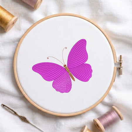 Vibrant purple butterfly machine embroidery design displayed inside a round wooden hoop on white fabric with thread spools and a needle.
