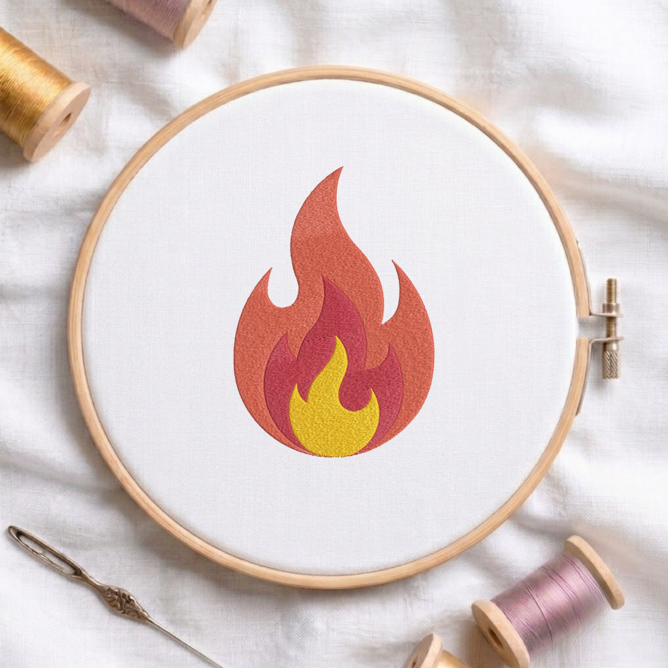 A circular embroidery hoop on white fabric holds a stylized flame design in orange, red, pink, and yellow. A needle and spools of thread are arranged around it on a white cloth background.