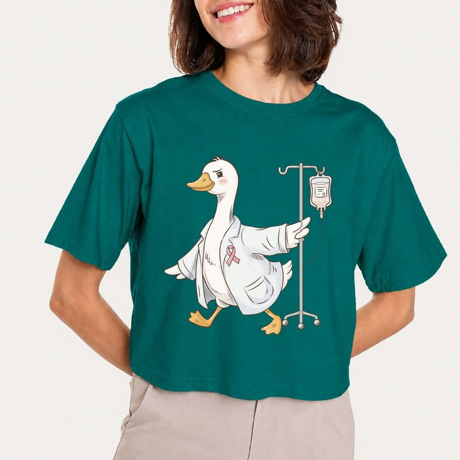 A woman wearing a dark teal cropped t-shirt with a graphic of a white goose in a medical coat next to an IV bag, paired with light beige pants, against a plain off-white background.