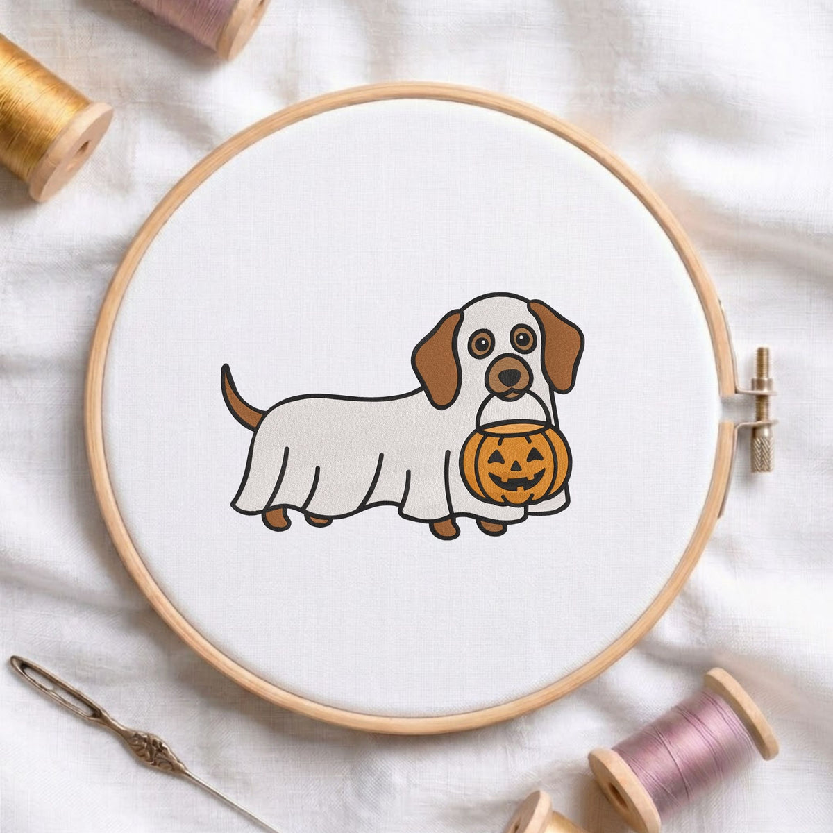 Ghost dachshund holding a pumpkin bucket machine embroidery design displayed in a wooden embroidery hoop on white fabric with thread spools and a needle