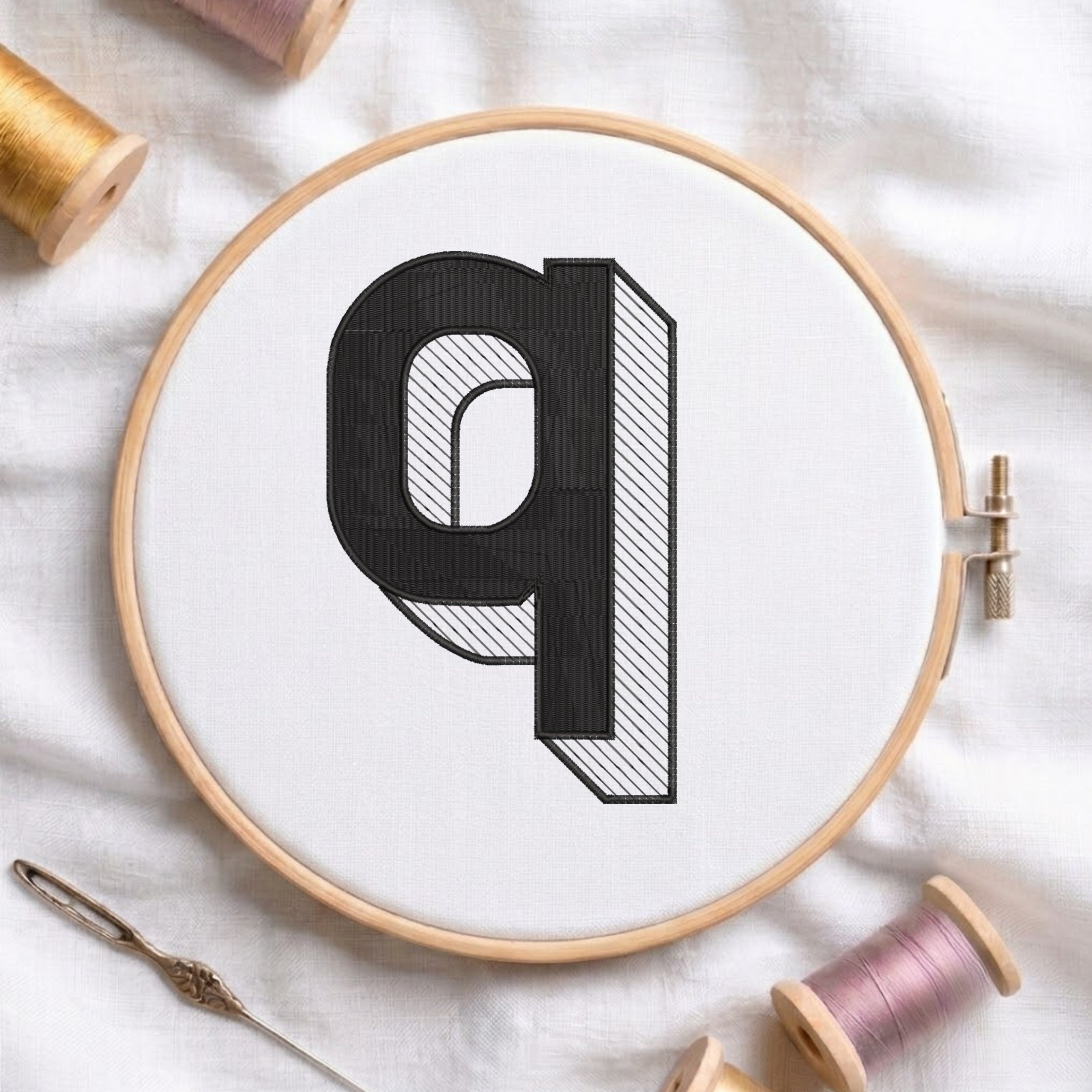 A wooden embroidery hoop holding white fabric with a large black letter 'q' design, surrounded by spools of thread and a needle on a white fabric background.