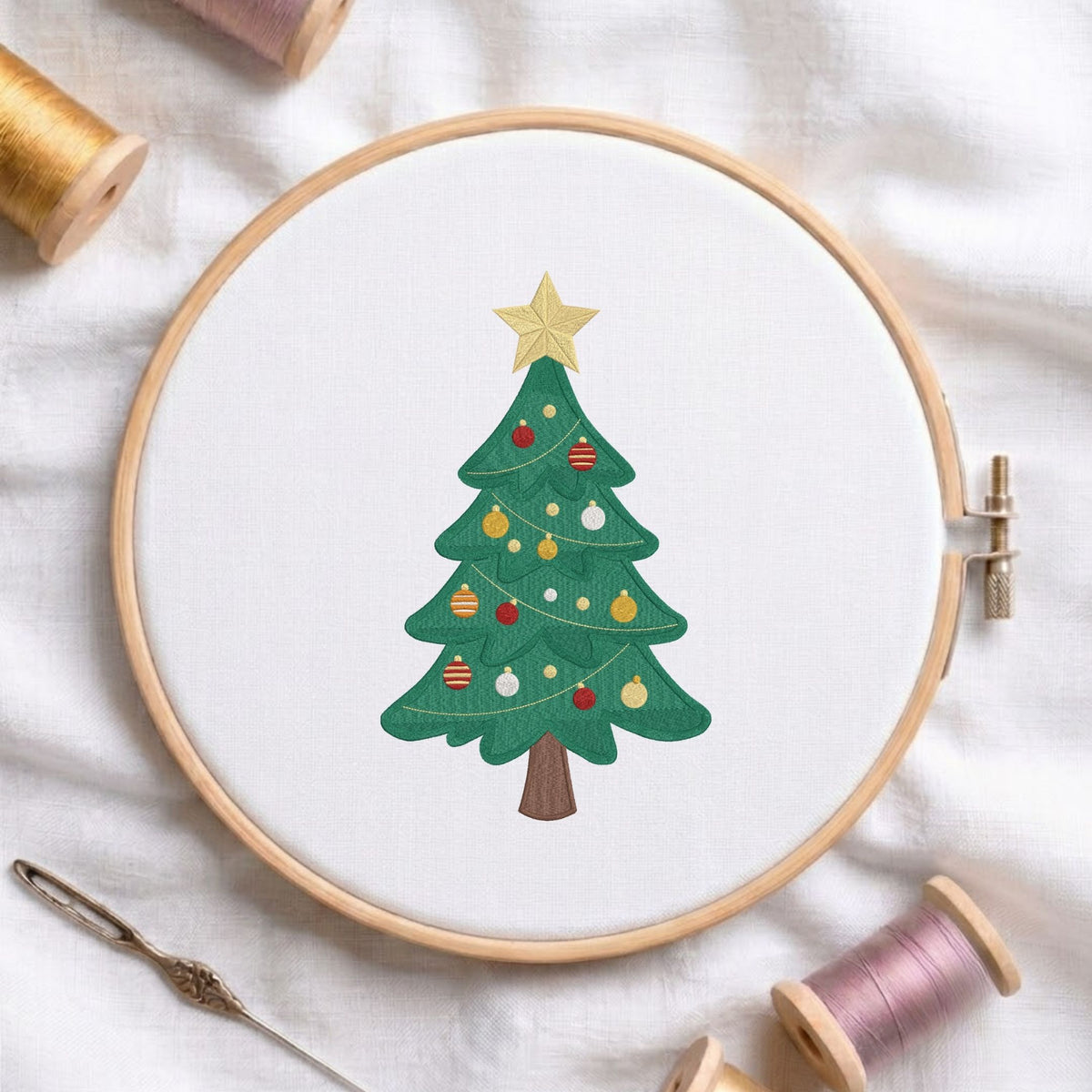 Classic decorated Christmas tree topped with a golden star machine embroidery design shown in a wooden embroidery hoop on white fabric with thread spools and a needle