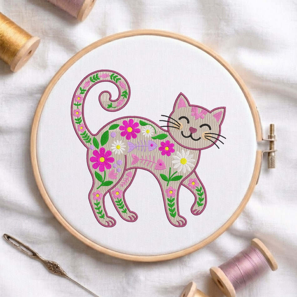 A wooden embroidery hoop holds a white fabric panel with a floral cat design stitched in pink, green, yellow, and white thread. The hoop is photographed on a white fabric background with spools of thread and a needle around it.