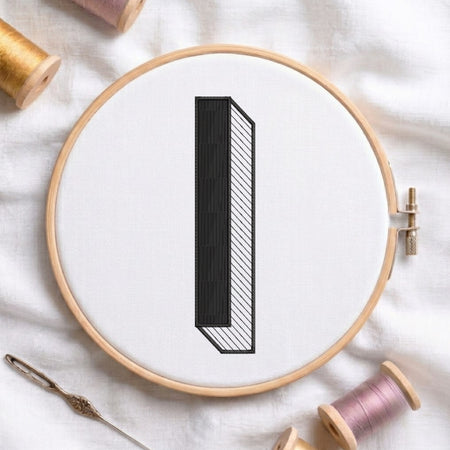 A wooden embroidery hoop holding white fabric with a minimalist black-and-white geometric letterform, styled on a white cloth with embroidery thread spools and a needle nearby.