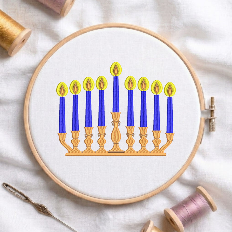 An embroidery hoop displays a completed design of a Hanukkah menorah with blue candles and gold holders on white fabric, surrounded by spools of thread and a needle.
