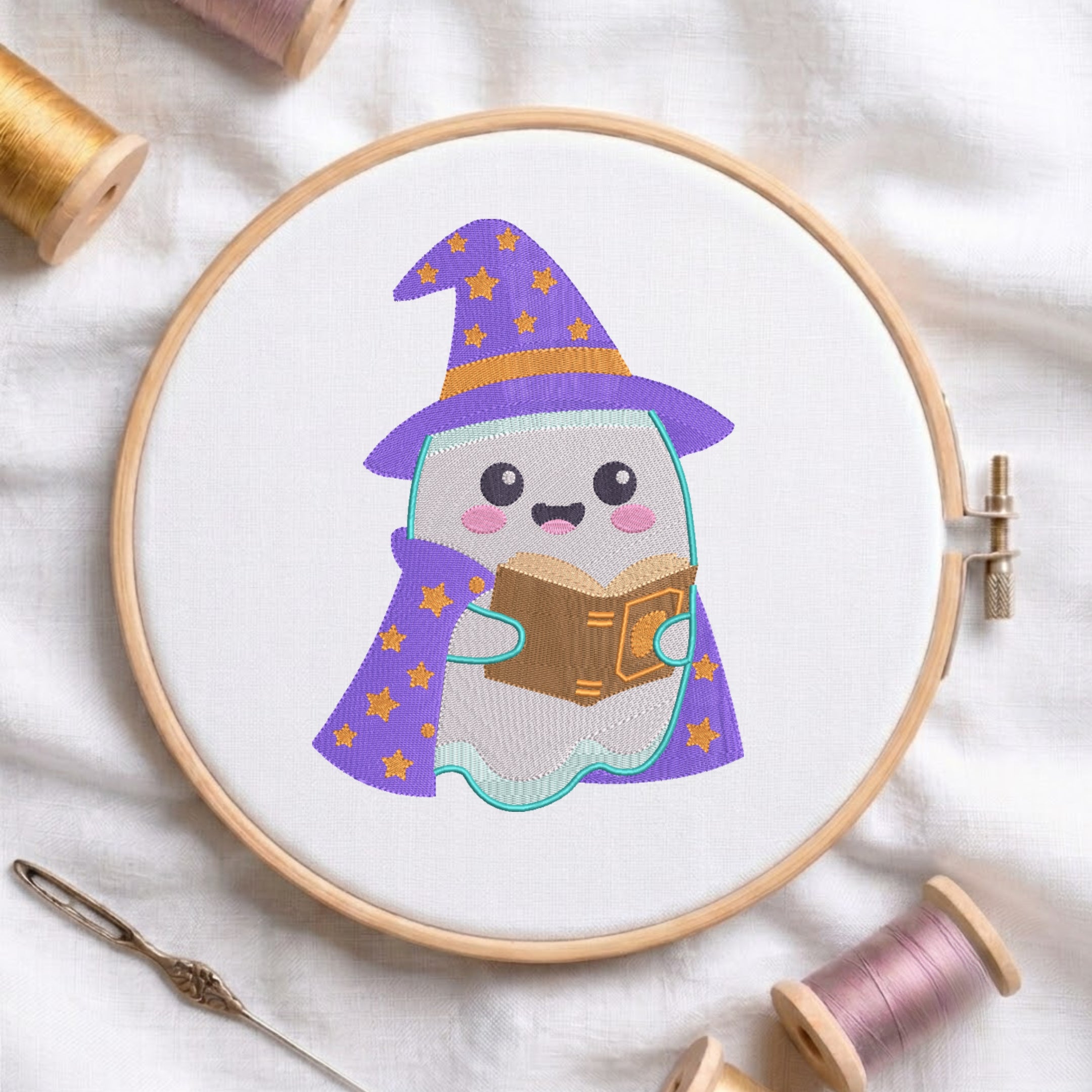 A circular embroidery hoop holding white fabric with a cute embroidered ghost character wearing a purple witch hat and matching cape with gold stars, reading a brown book. The hoop is styled on a white fabric background with spools of thread and a needle nearby.