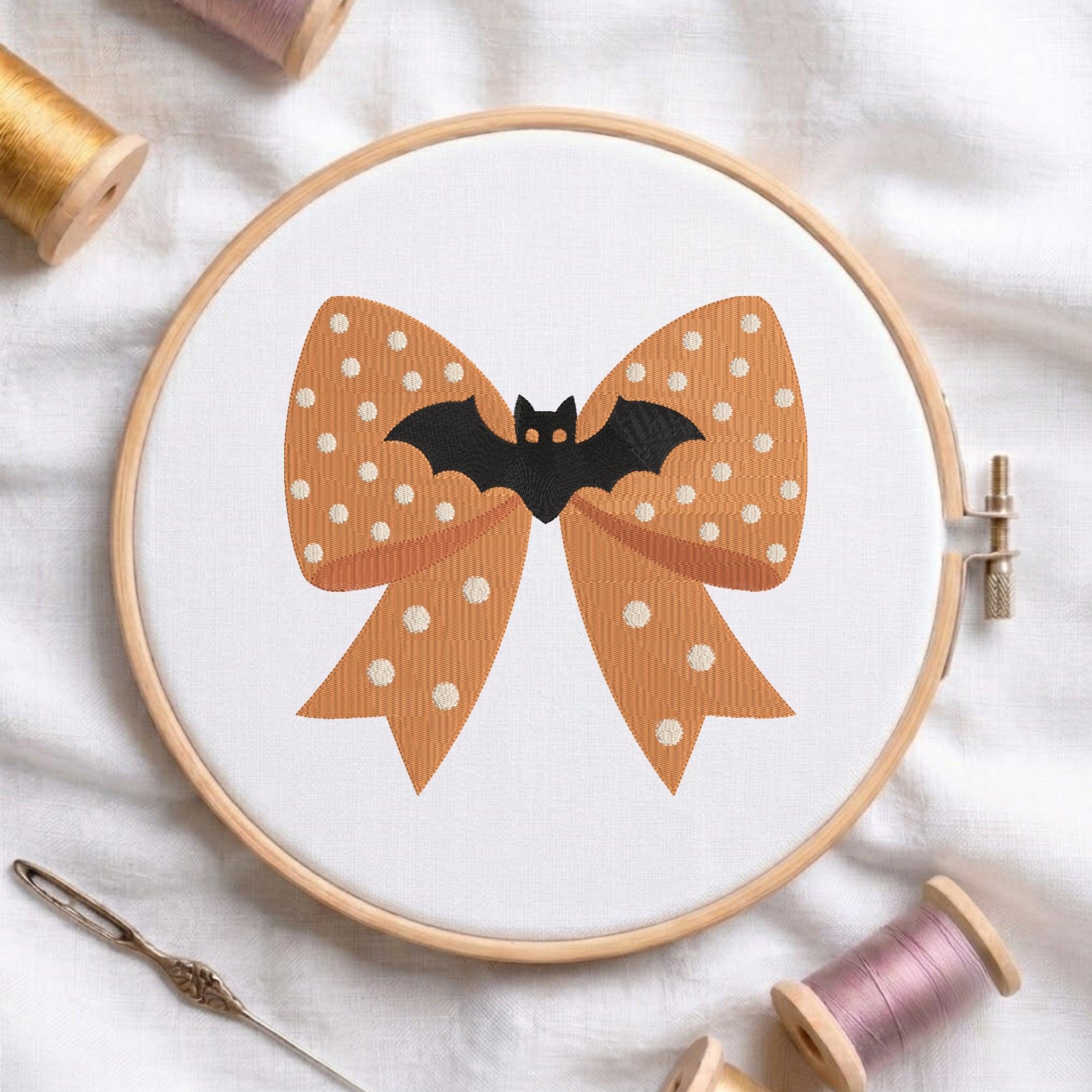 white fabric stretched in a wooden embroidery hoop, featuring an embroidered design of an orange polka-dot bow centered with a cute black bat.