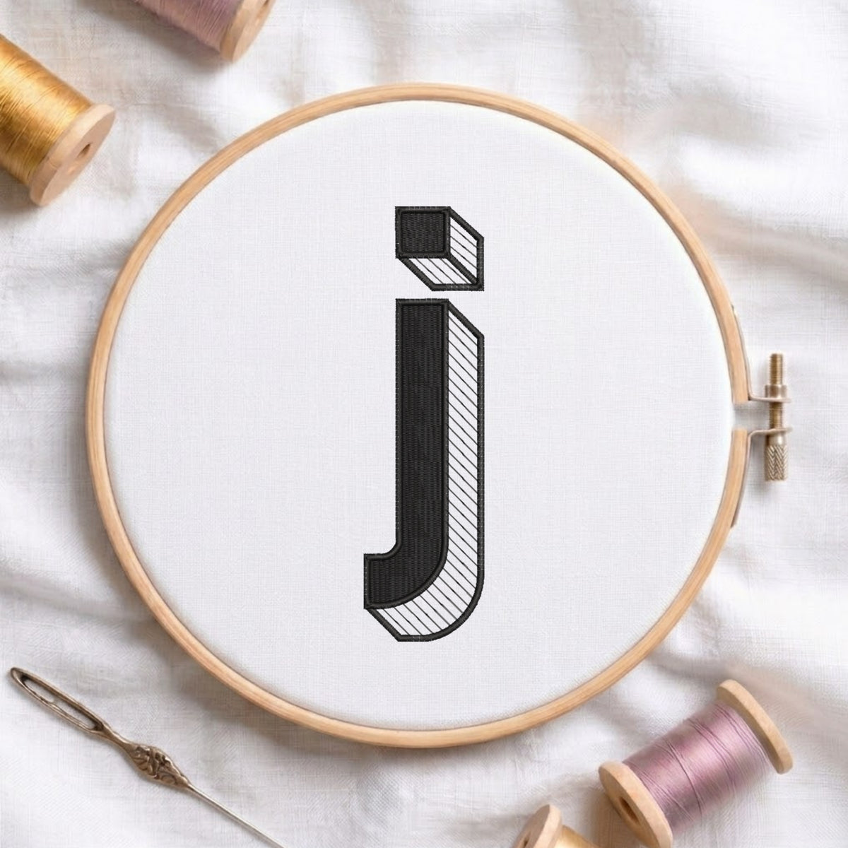 A wooden embroidery hoop holding white fabric with a large black embroidered letter 'j' in a stylized block font. The hoop is photographed on a white fabric surface with spools of thread and a needle around it.