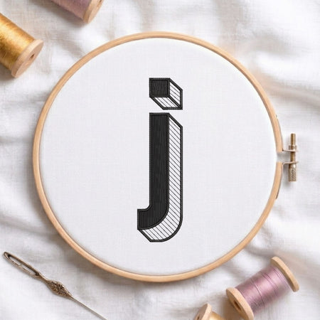 A wooden embroidery hoop holding white fabric with a large black embroidered letter 'j' in a stylized block font. The hoop is photographed on a white fabric surface with spools of thread and a needle around it.