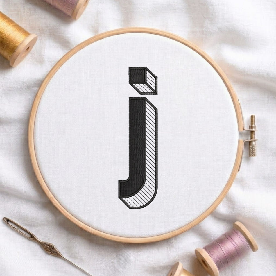 A wooden embroidery hoop holding white fabric with a large black embroidered letter 'j' in a stylized block font. The hoop is photographed on a white fabric surface with spools of thread and a needle around it.