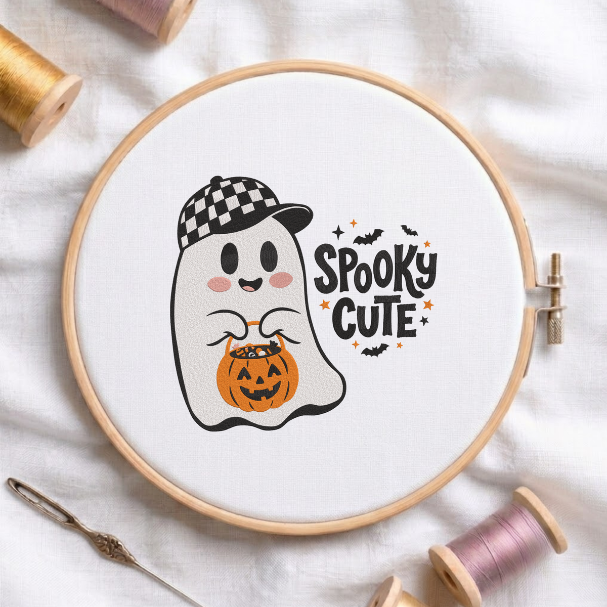 "Spooky Cute" ghost wearing a checkered baseball cap and holding a pumpkin candy bucket machine embroidery design shown in a wooden embroidery hoop on white fabric with thread spools and a needle