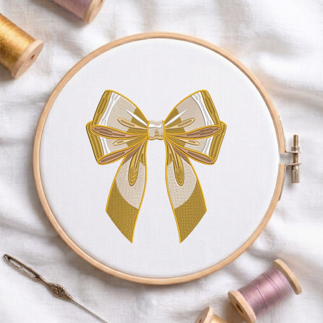 A circular embroidery hoop on white fabric, featuring a bow design in gold and cream thread. Spools of thread and a needle are arranged around the hoop on a white fabric background.