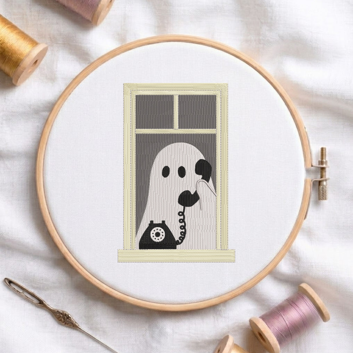 A circular embroidery hoop on white fabric, showing a stitched design of a ghost talking on a rotary telephone. Spools of thread and a needle are arranged around the hoop.