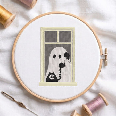 A circular embroidery hoop on white fabric, showing a stitched design of a ghost talking on a rotary telephone. Spools of thread and a needle are arranged around the hoop.