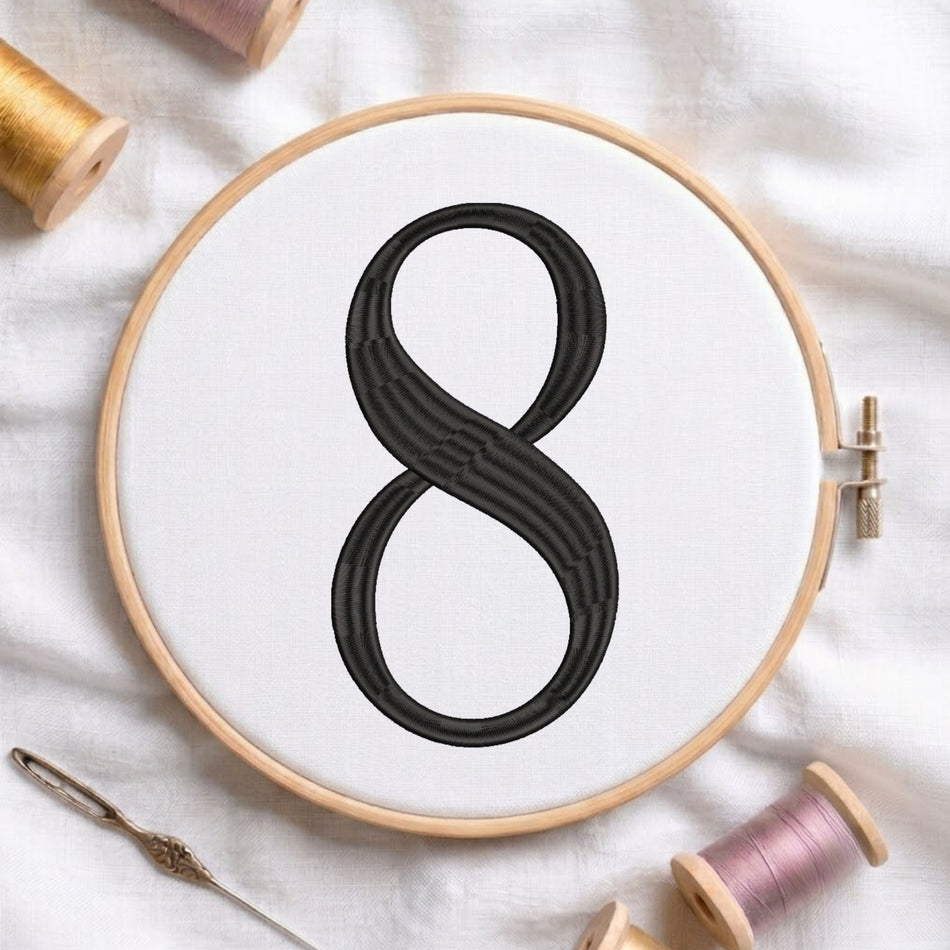 A wooden embroidery hoop holds white fabric with a large black stitched infinity symbol. The hoop is photographed on a white cloth surface with spools of thread and a needle nearby.