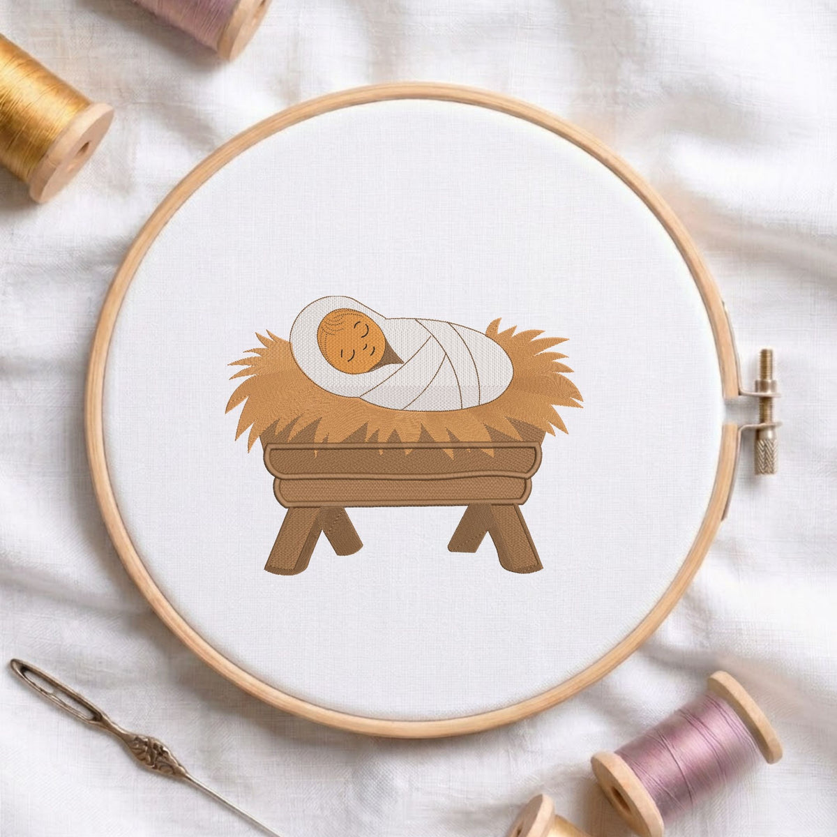 Baby Jesus in a manger machine embroidery design featuring a swaddled infant resting in straw inside a wooden crib, displayed inside a round wooden hoop on white linen fabric alongside thread spools and a needle.