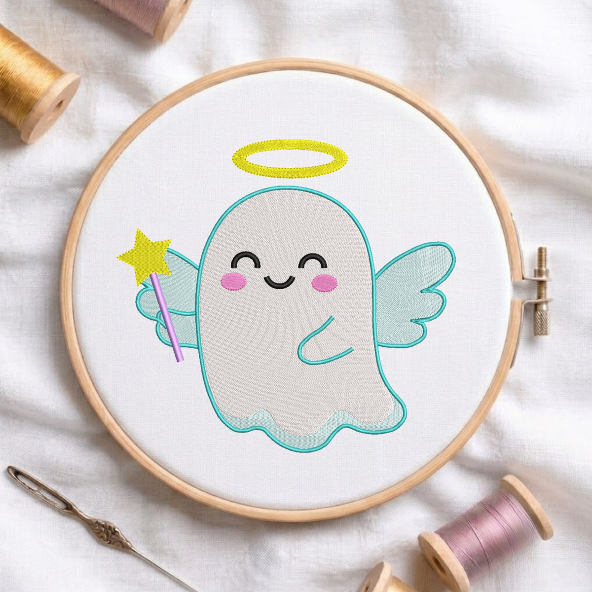 A circular embroidery hoop holding white fabric with a cute ghost angel design. The design features a light gray ghost with light blue wings, a yellow halo, pink cheeks, and a yellow wand.