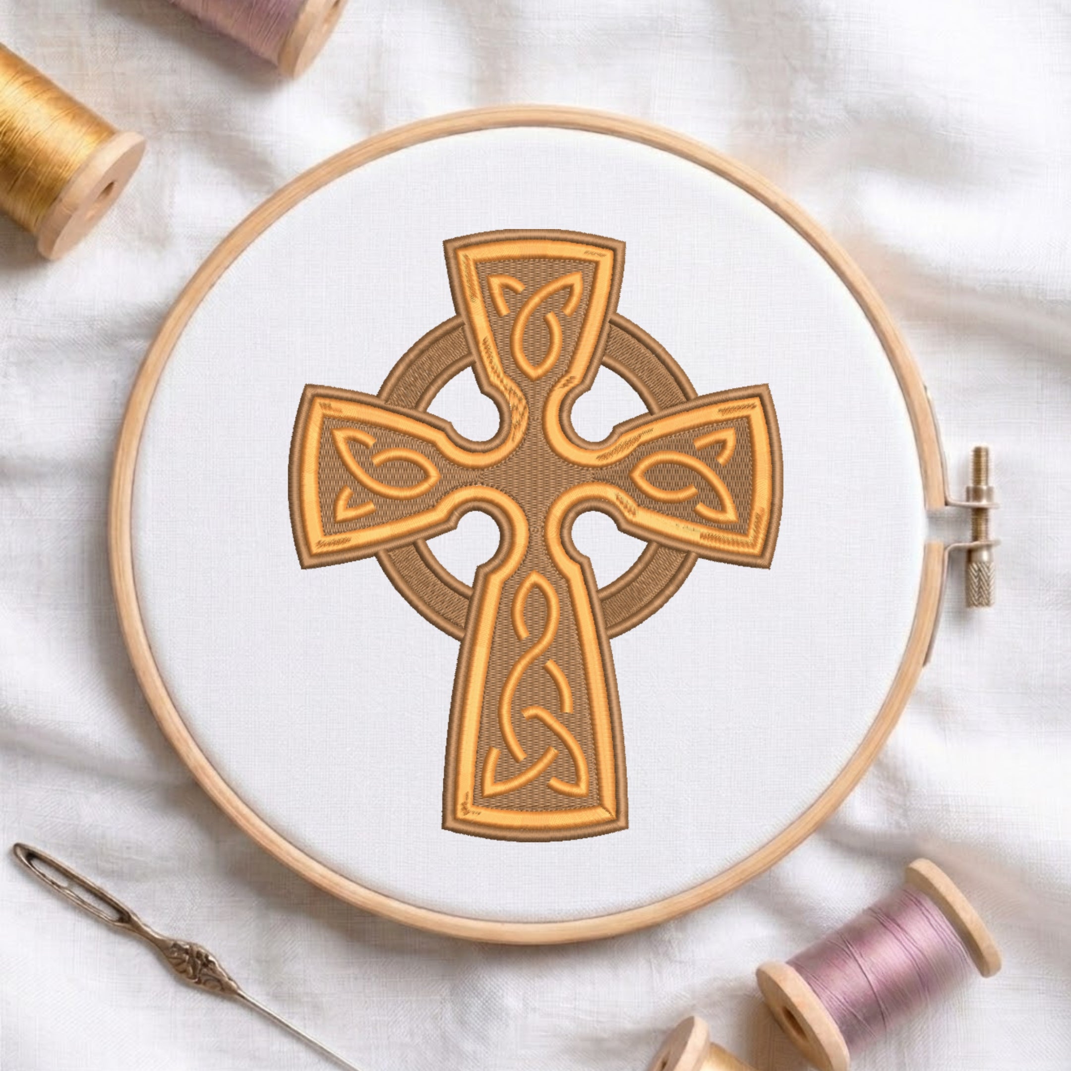 Intricate gold and brown Celtic cross with knotwork embroidered on white fabric inside a round wooden hoop with thread spools.