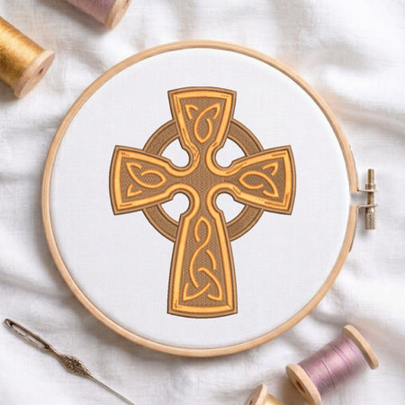 Intricate gold and brown Celtic cross with knotwork embroidered on white fabric inside a round wooden hoop with thread spools.