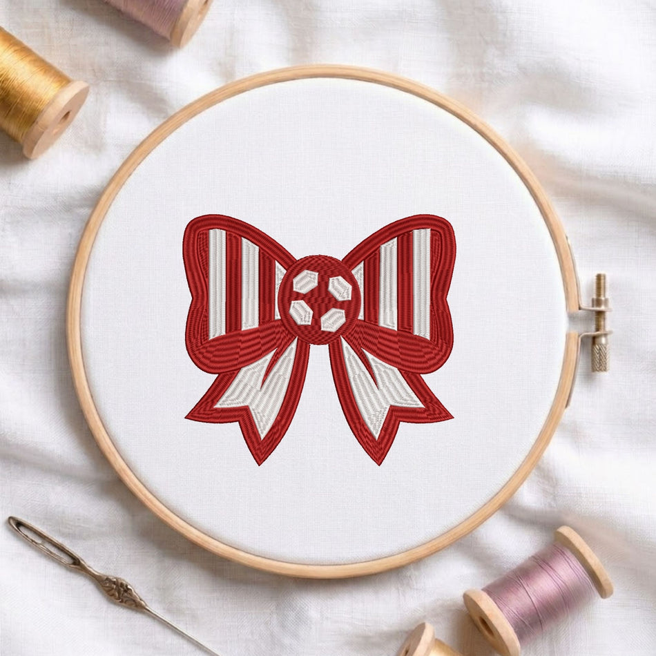 Red and white striped bow machine embroidery design featuring a soccer ball center, displayed inside a round wooden hoop on white fabric with thread spools and a needle.