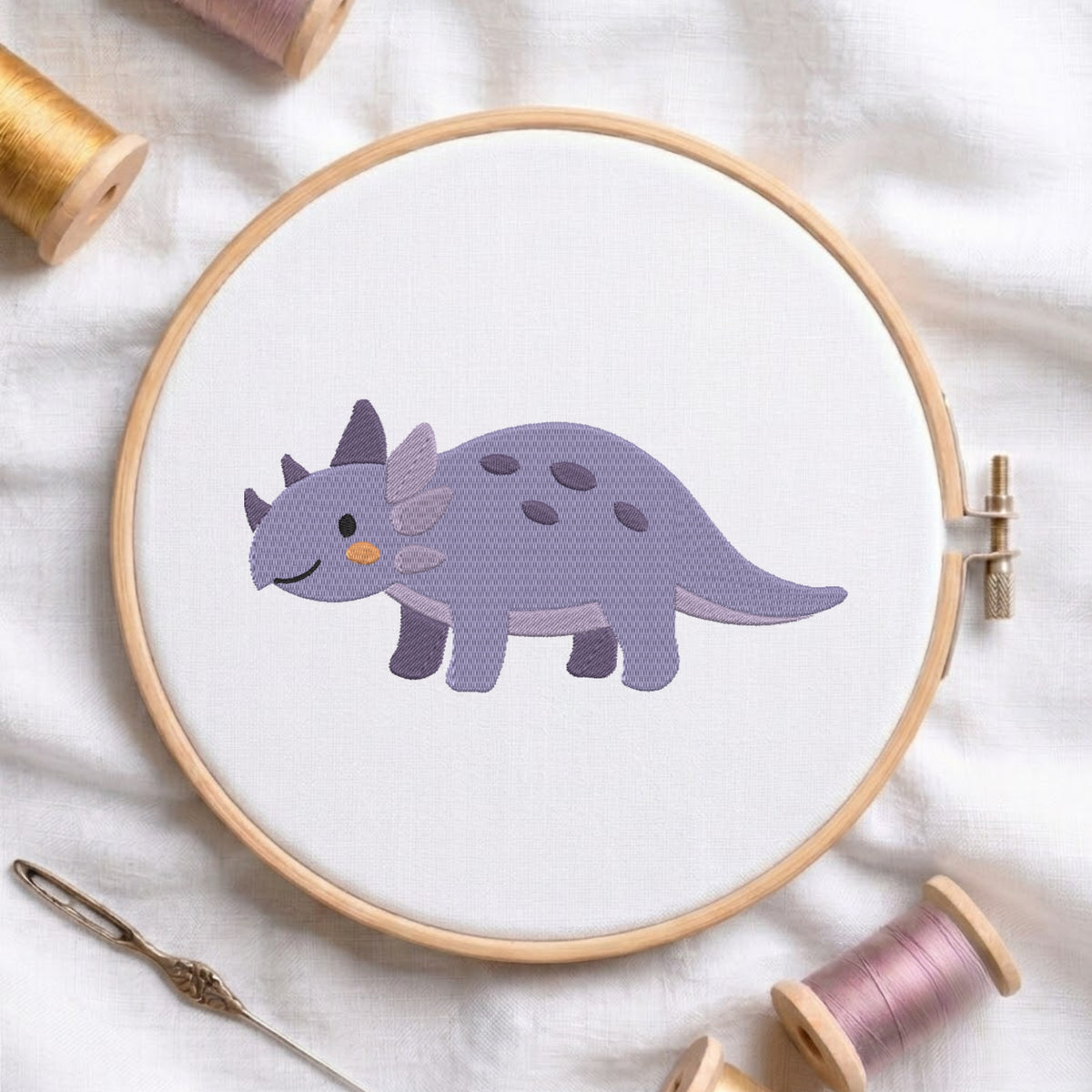 A wooden embroidery hoop holds a white fabric panel with a stitched cartoon dinosaur in lavender and gray. The hoop is photographed on a white fabric surface with spools of thread and a needle around it.